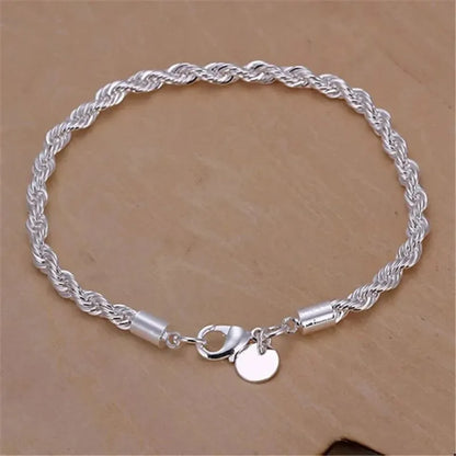 Silver twisted rope chain bracelet laid flat in an oval on a tan wooden surface, showing the lobster clasp and round tag charm.