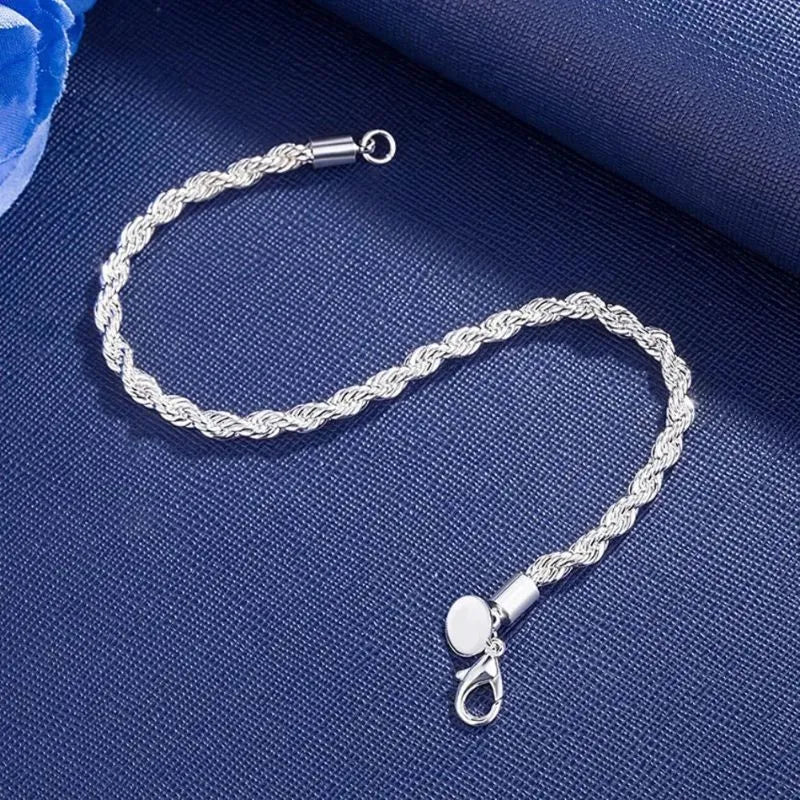 Silver twisted rope chain bracelet laid in an S-curve on a navy fabric backdrop with a blue rose prop, showing both clasp ends and the round tag charm.