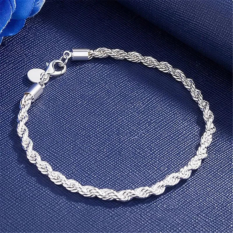 Silver twisted rope chain bracelet laid in a circle on a navy fabric backdrop with a blue rose prop, showing the lobster clasp and round tag charm.
