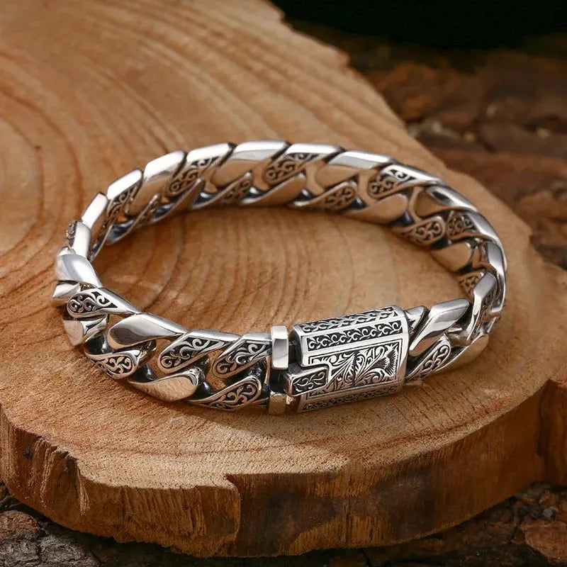 Engraved silver Cuban-link bracelet laid on a raw wood slab, showing the vintage vine-and-scroll pattern on each link.