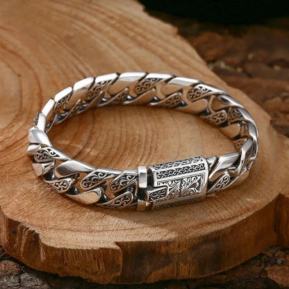 Engraved silver Cuban-link bracelet laid on a raw wood slab, showing the vintage vine-and-scroll pattern on each link.