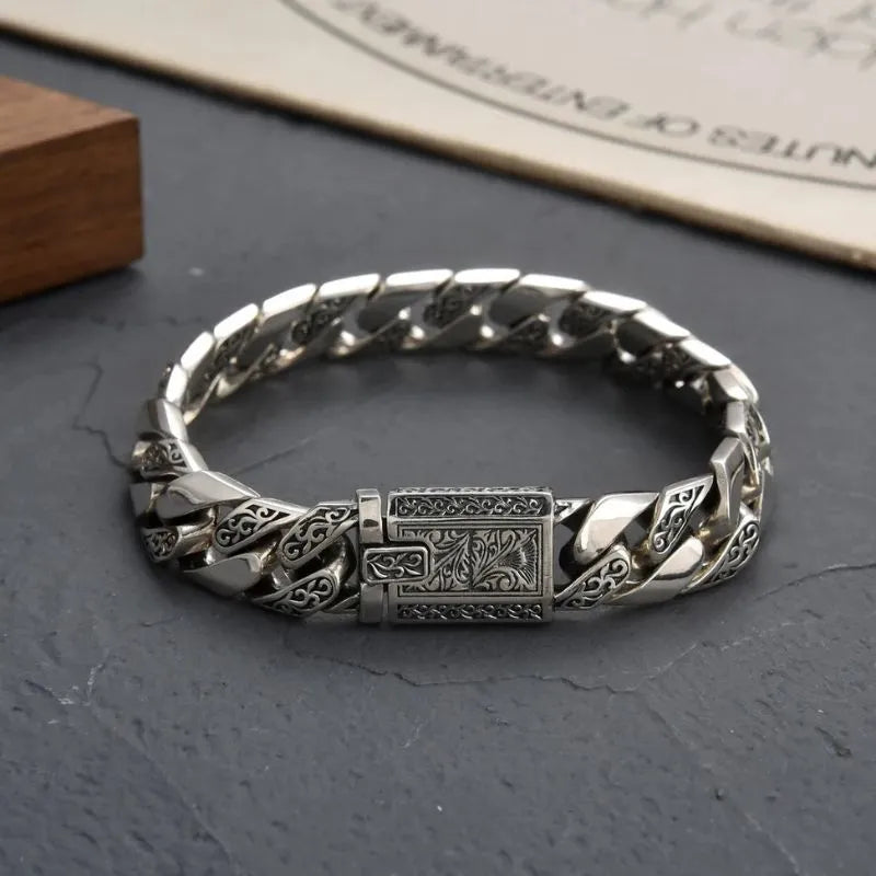 Engraved silver Cuban-link bracelet laid in an oval on a dark grey stone surface, with a wooden block prop and a partially visible printed page in the background.