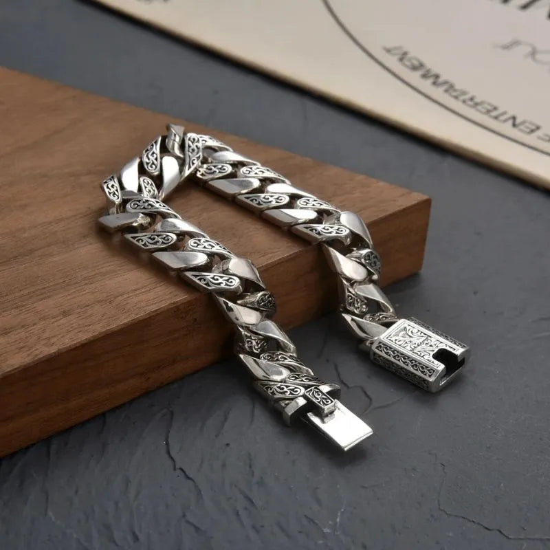 Engraved silver Cuban-link bracelet laid open and diagonal over a wooden block on a dark stone surface, with a certificate-style paper in the background.