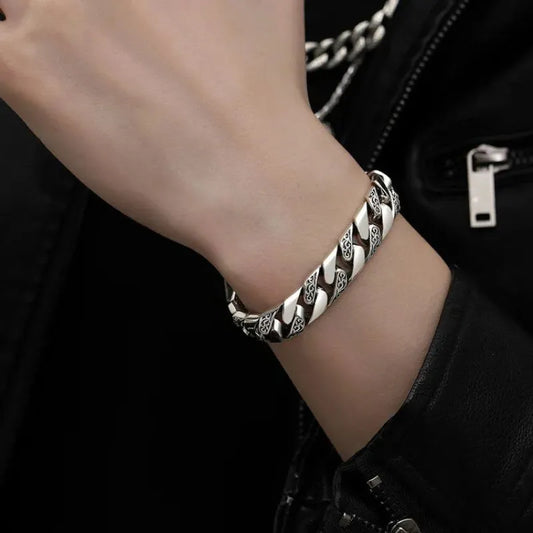 Engraved silver Cuban-link bracelet worn on the wrist, paired with a black leather jacket and a layered silver chain necklace.