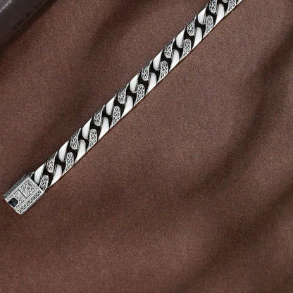 Close-up of the engraved Cuban-link bracelet laid diagonally on a brown fabric surface.