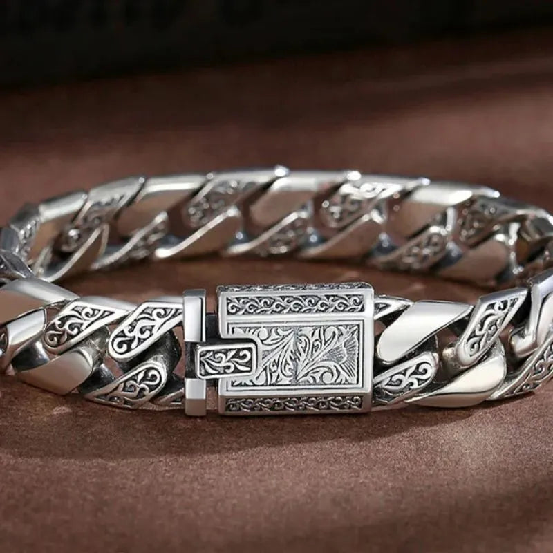 Engraved silver Cuban-link bracelet laid in an oval on a brown leather-like surface, clasp facing forward.