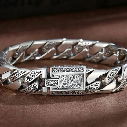 Engraved silver Cuban-link bracelet laid in an oval on a brown leather-like surface, clasp facing forward.