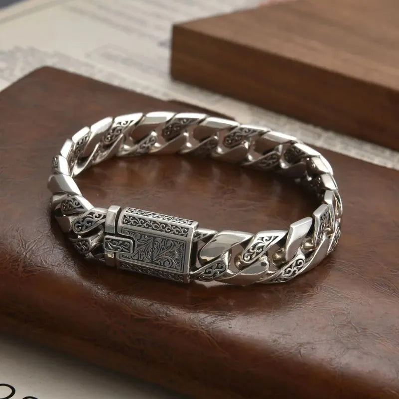 Engraved silver Cuban-link bracelet on a brown leather-textured surface, with a paper document and wooden block prop in the background.
