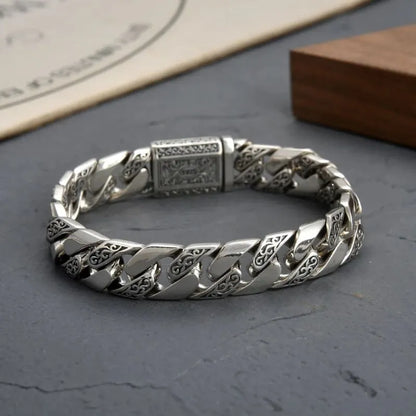 Close-up of the engraved Cuban-link bracelet's clasp, laid on a dark grey stone slab with paper and wooden block props in the background.
