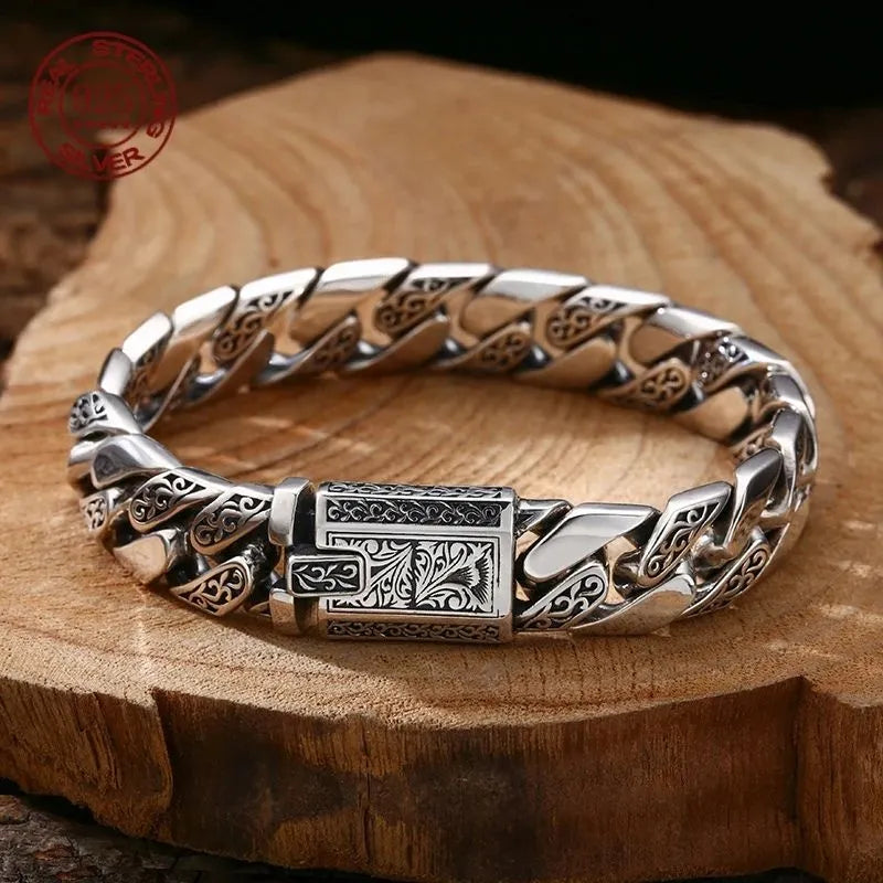 Engraved silver Cuban-link bracelet with an ornate rectangular clasp, laid on a raw wood slab with a red sterling silver hallmark stamp visible in the corner.