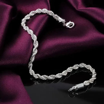 Silver twisted rope chain bracelet draped diagonally on dark wine-colored satin fabric, with a reflective black surface below.