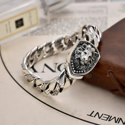 Silver Cuban-link bracelet with a lion-head shield centerpiece, laid on a marble surface beside a leather journal and printed page.
