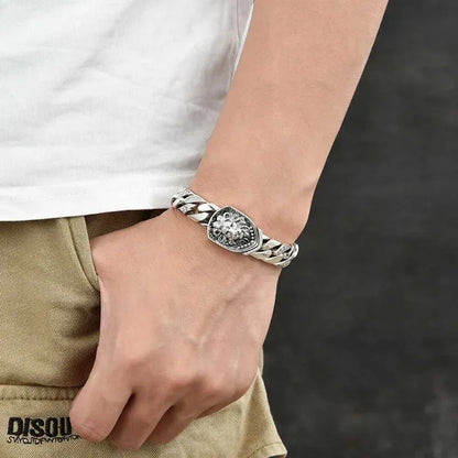 Silver Cuban-link bracelet with a lion-head shield centerpiece worn on the wrist, paired with a casual white t-shirt and khaki pants.