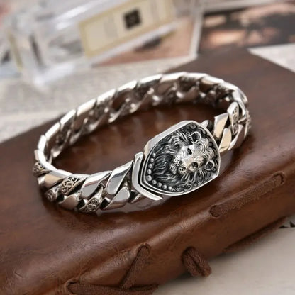 Silver Cuban-link bracelet with a sculpted lion-head shield centerpiece, laid on a brown leather journal with blurred paper props in the background.