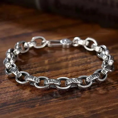 Silver mantra-engraved link bracelet laid in an oval on a wooden surface, with a dark rounded prop in the background.