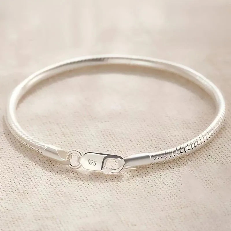 Silver snake chain bracelet laid in a circle on beige textured fabric, with a '925' hallmark stamped on the clasp.