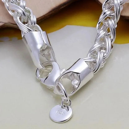 Close-up of the silver wheat-chain bracelet's clasp with a round '925' tag charm, propped against a leather-like surface.