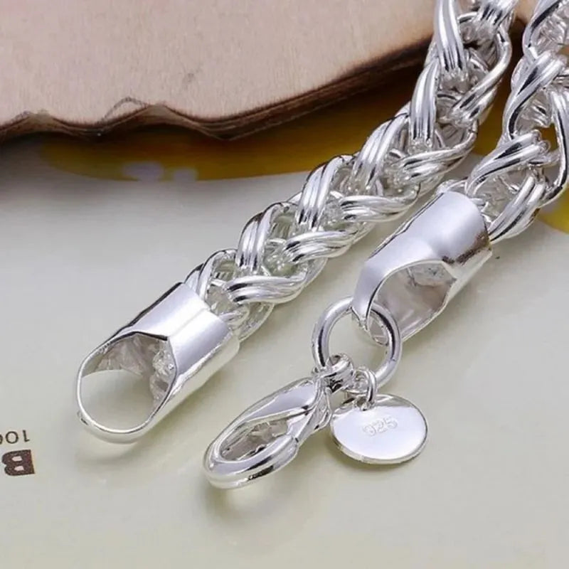 Close-up of the silver wheat-chain bracelet's clasp end and round '925' tag charm, propped against a leather-like surface.
