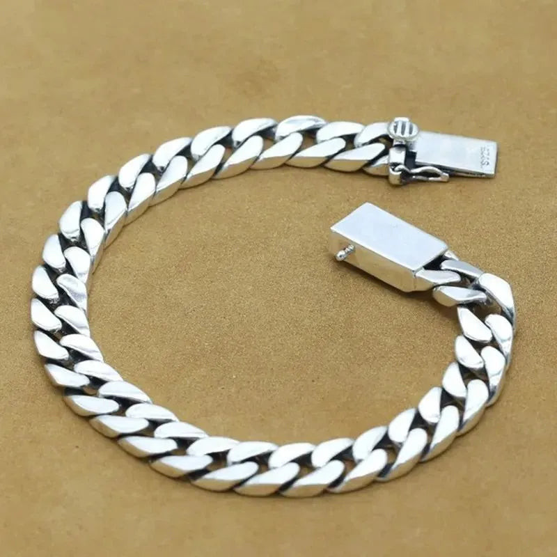 Silver Cuban-link bracelet shown open and unclasped, laid in a circle on a tan suede surface.
