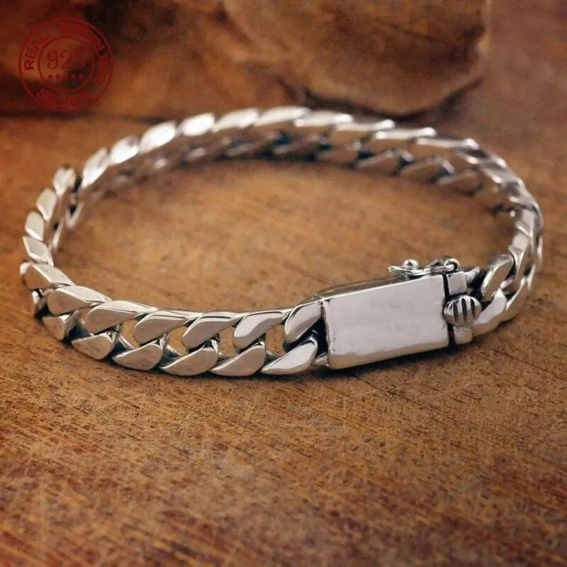 Silver Cuban-link bracelet laid in an oval on a textured wooden surface, with a stone prop and red sterling silver hallmark stamp visible.