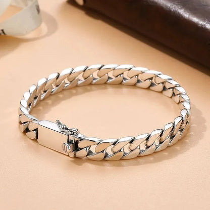 Silver Cuban-link bracelet laid in an oval on a peach-toned surface, with a dark leather pouch in the background.