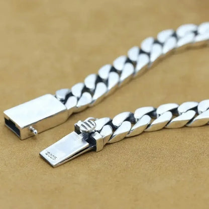 Silver Cuban-link bracelet shown open and unclasped, close-up of the 'S925'-stamped clasp mechanism on a tan suede surface.