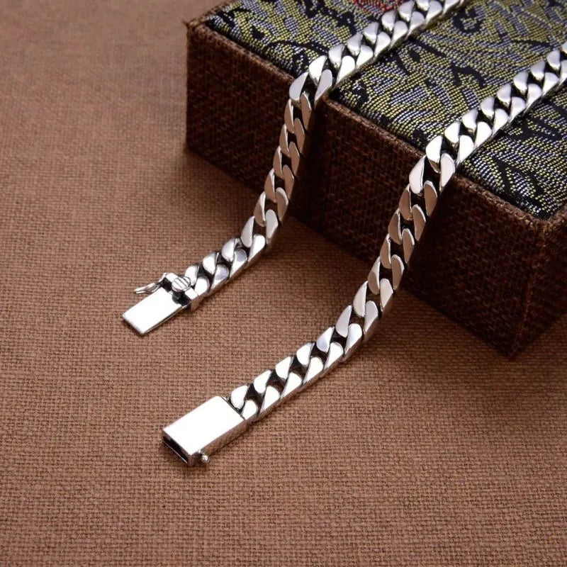Silver Cuban-link bracelet shown open and unclasped, draped over a decorative patterned box, on a brown woven fabric surface.