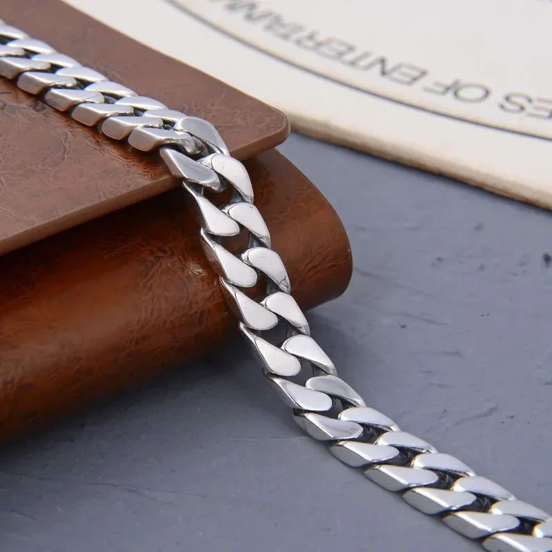 Silver Cuban-link bracelet draped over a rounded brown leather-like prop, on a dark grey stone surface with a printed page in the background.