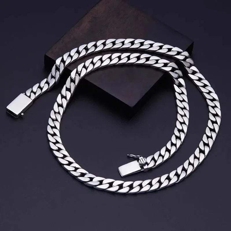 Long silver Cuban-link chain coiled in an S-curve on a dark surface, with a wooden block prop and the clasp shown open.