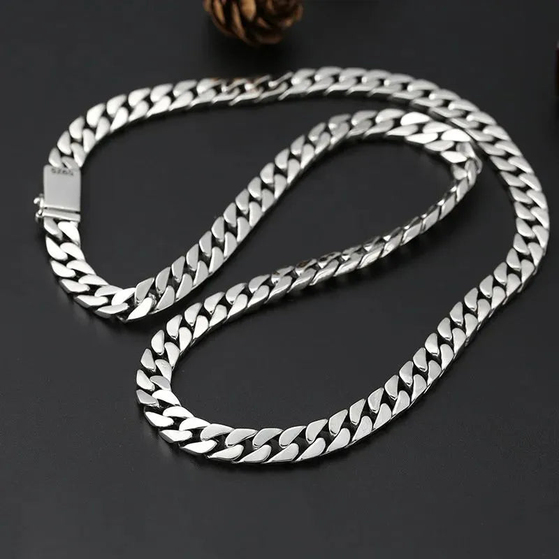 Long silver Cuban-link chain coiled on a dark surface, with a pinecone-shaped prop and an 'S925' stamp visible on the clasp.