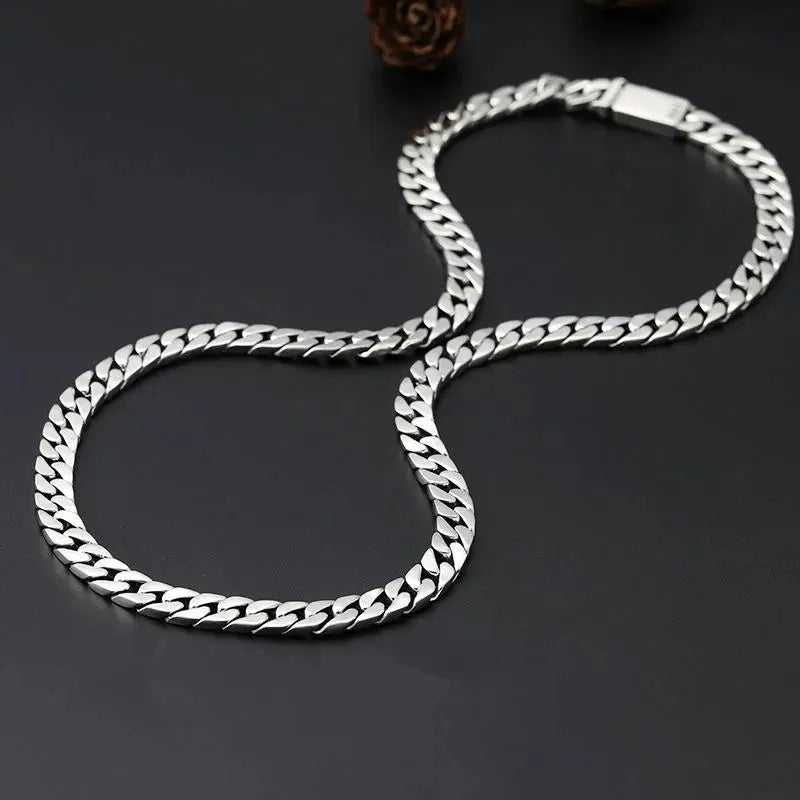 Long silver Cuban-link chain coiled in a figure-eight shape on a dark surface, with a pinecone-shaped prop.