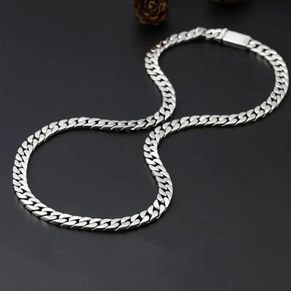 Long silver Cuban-link chain coiled in a figure-eight shape on a dark surface, with a pinecone-shaped prop.