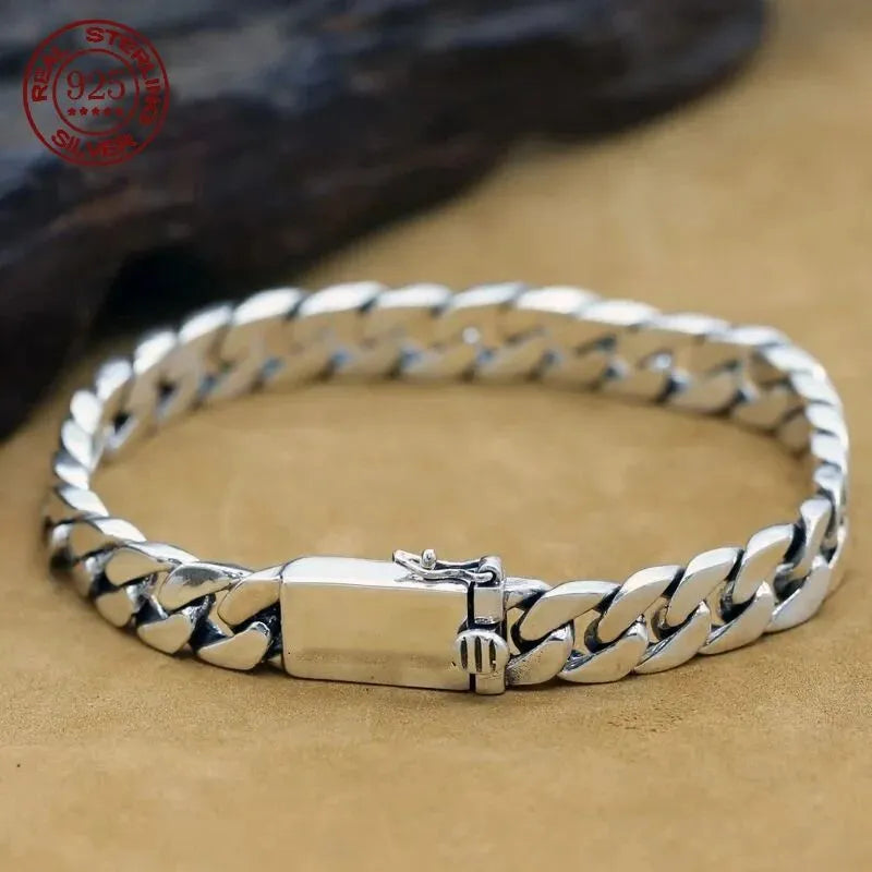 Silver Cuban-link bracelet with a rectangular box clasp, laid on a tan suede surface with a dark rock prop and a red 'Real Sterling Silver 925' hallmark stamp in the corner.