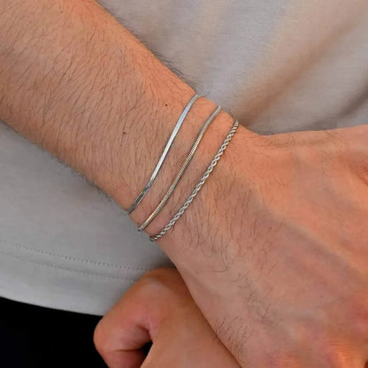 Three silver bracelets — flat blade, snake, and twisted rope chains — stacked on the wrist, against a grey shirt background.