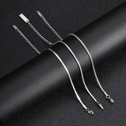 Three silver chains — flat blade, snake, and twisted rope — laid out separately with clasps and extension chains visible, draped over a curved black leather surface.