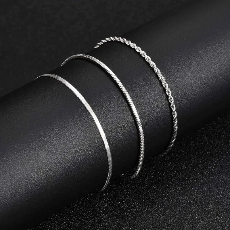 Three silver chains — flat blade, snake, and twisted rope — coiled together side by side on a black leather surface, showing the texture and width comparison.