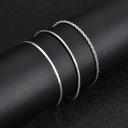 Three silver chains — flat blade, snake, and twisted rope — coiled together side by side on a black leather surface, showing the texture and width comparison.