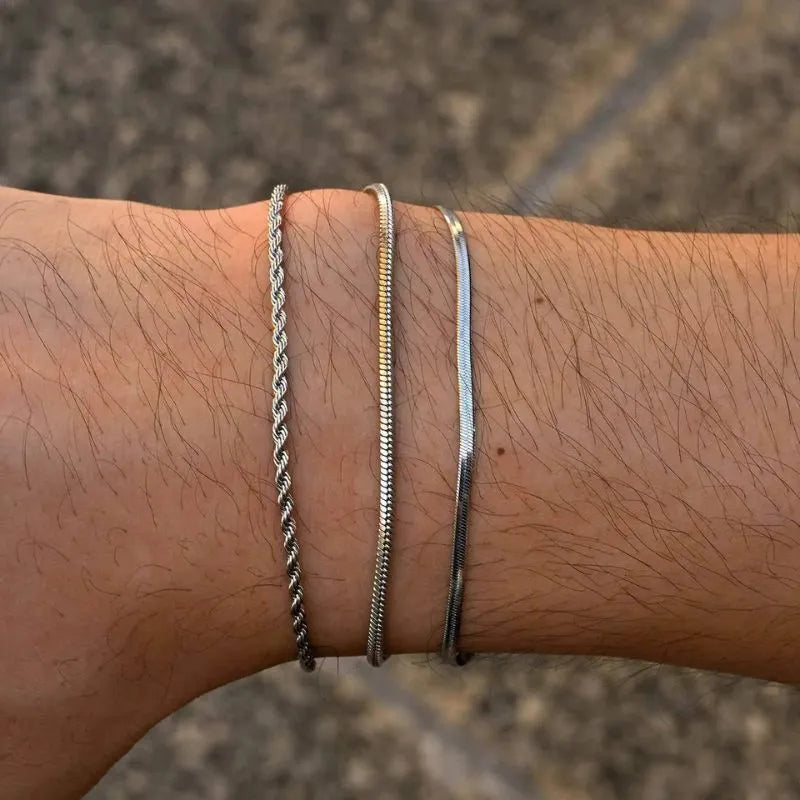 Three silver bracelets — twisted rope, snake, and flat blade chains — stacked on the wrist, close-up outdoors against a stone tile background.