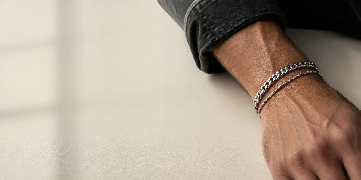 Man's wrist wearing a stainless steel curb chain bracelet and a slim snake chain bracelet, resting on an off-white surface in natural window light